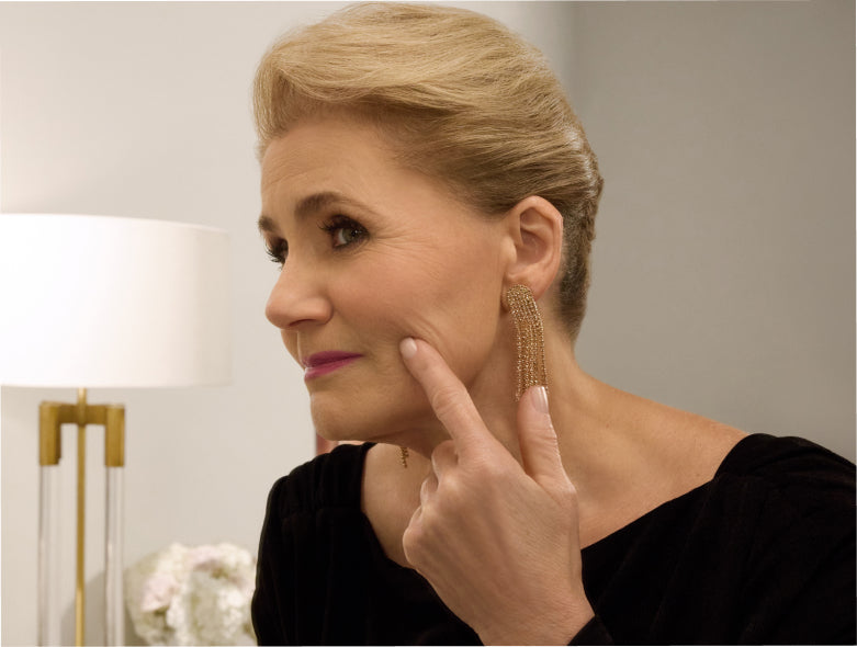 Woman wearing gold hoop earrings, looking at her facial lines in the mirror.